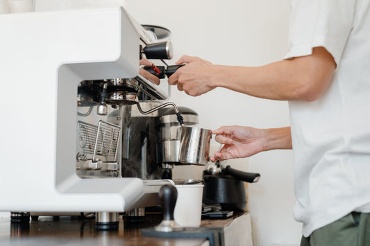 Barista steaming milk in a stainless steel pitcher at an espresso machine to create microfoam.